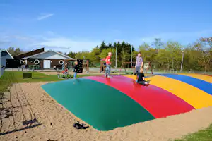 Buntes Sandspielfeld auf dem Campingplatz, daneben Gebäude und Spielgeräte, im Hintergrund Bäume und ein blauer Himmel.