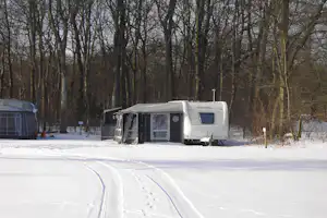 Wohnwagen und Vorzelt auf einem Campingplatz im verschneiten Wald, mit Fahrspur im Schnee und Bäumen im Hintergrund.