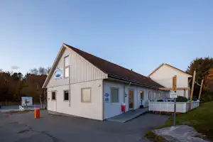 Campinggebäude mit Rezeption und Café-Schildern, weißem Holzbau, Fenstern und Eingangstreppe vor Asphaltweg und Bäumen.