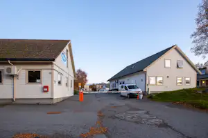 Campingplatz-Gebäude mit Stellplätzen und Wohnwagen vor weißen Häusern, Asphaltweg und Bäumen am Rand.
