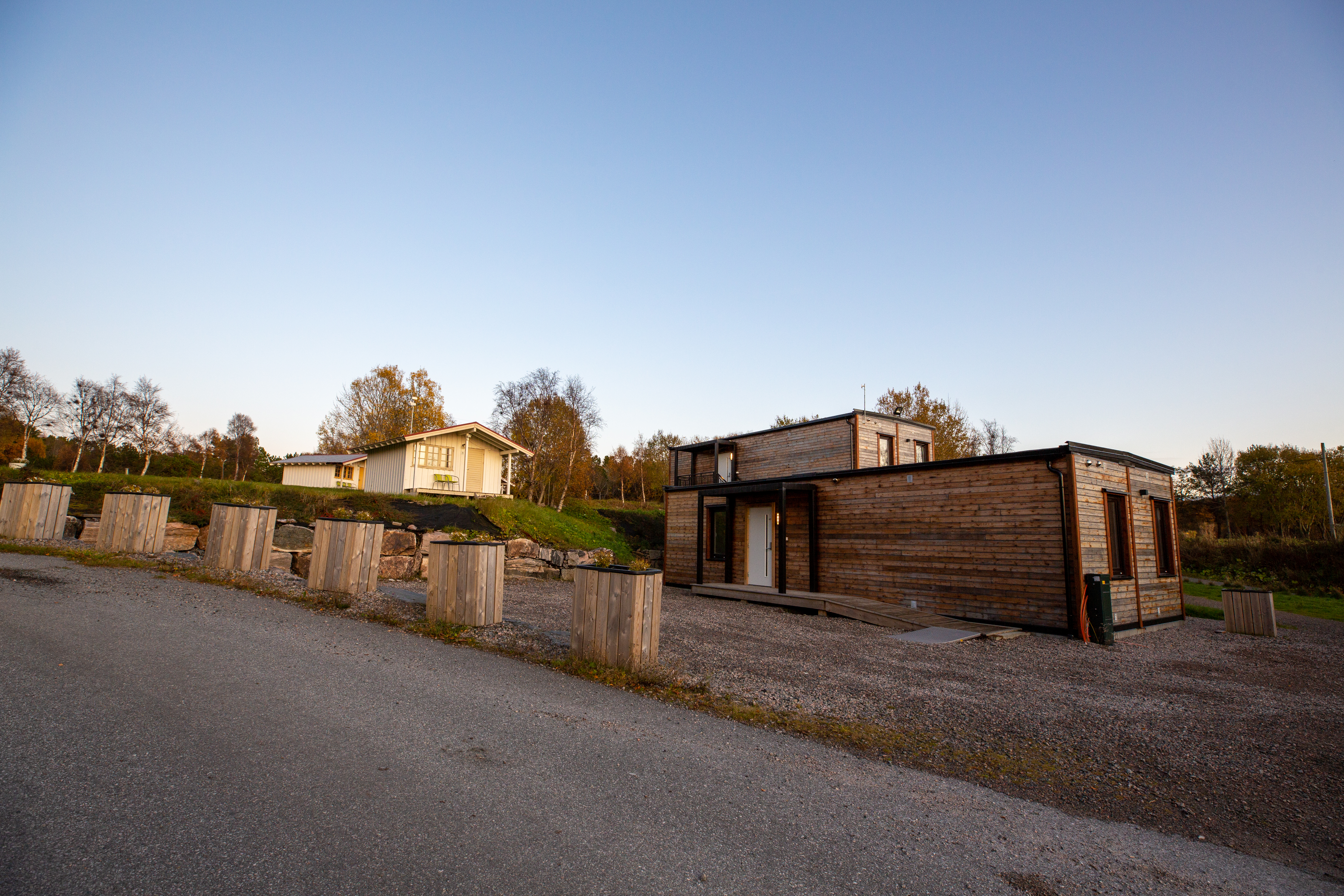 Holzunterkunft und Stellplatz am Campingplatz, mit Schotterweg, Zaun und Bäumen im Hintergrund.