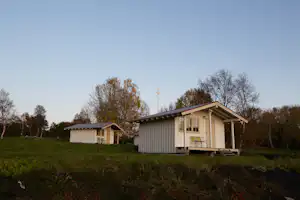Zwei kleine Holz-Campinghütten auf einer Wiese, mit Veranda und Sitzbank, im Hintergrund Bäume und ein Schuppen.