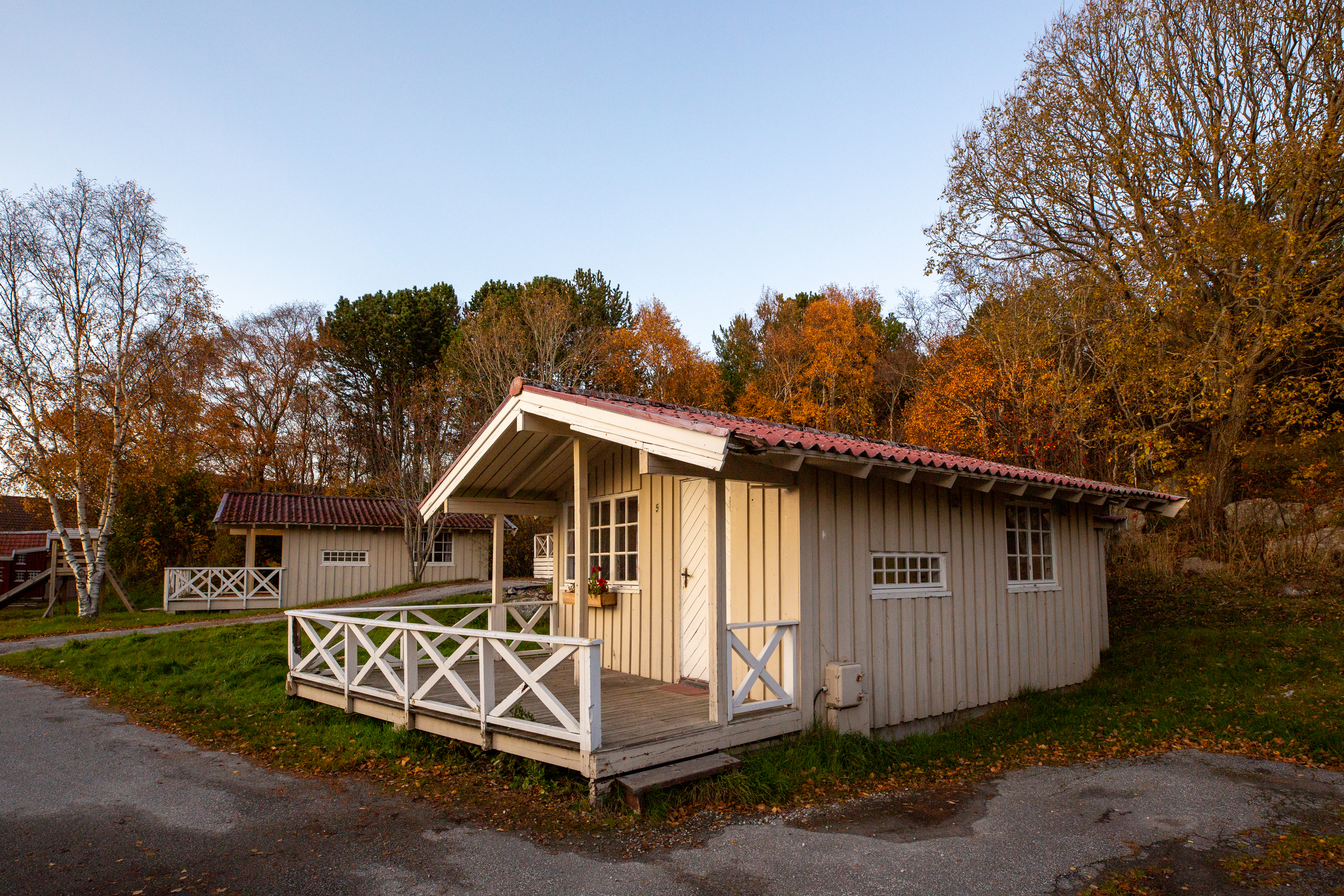 Holz-Campinghütte mit Veranda, Sitzbank und Geländer auf einem Stellplatz, umgeben von Bäumen im Herbst.