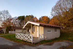 Holz-Campinghütte mit Veranda, Sitzbank und Geländer auf einem Stellplatz, umgeben von Bäumen im Herbst.