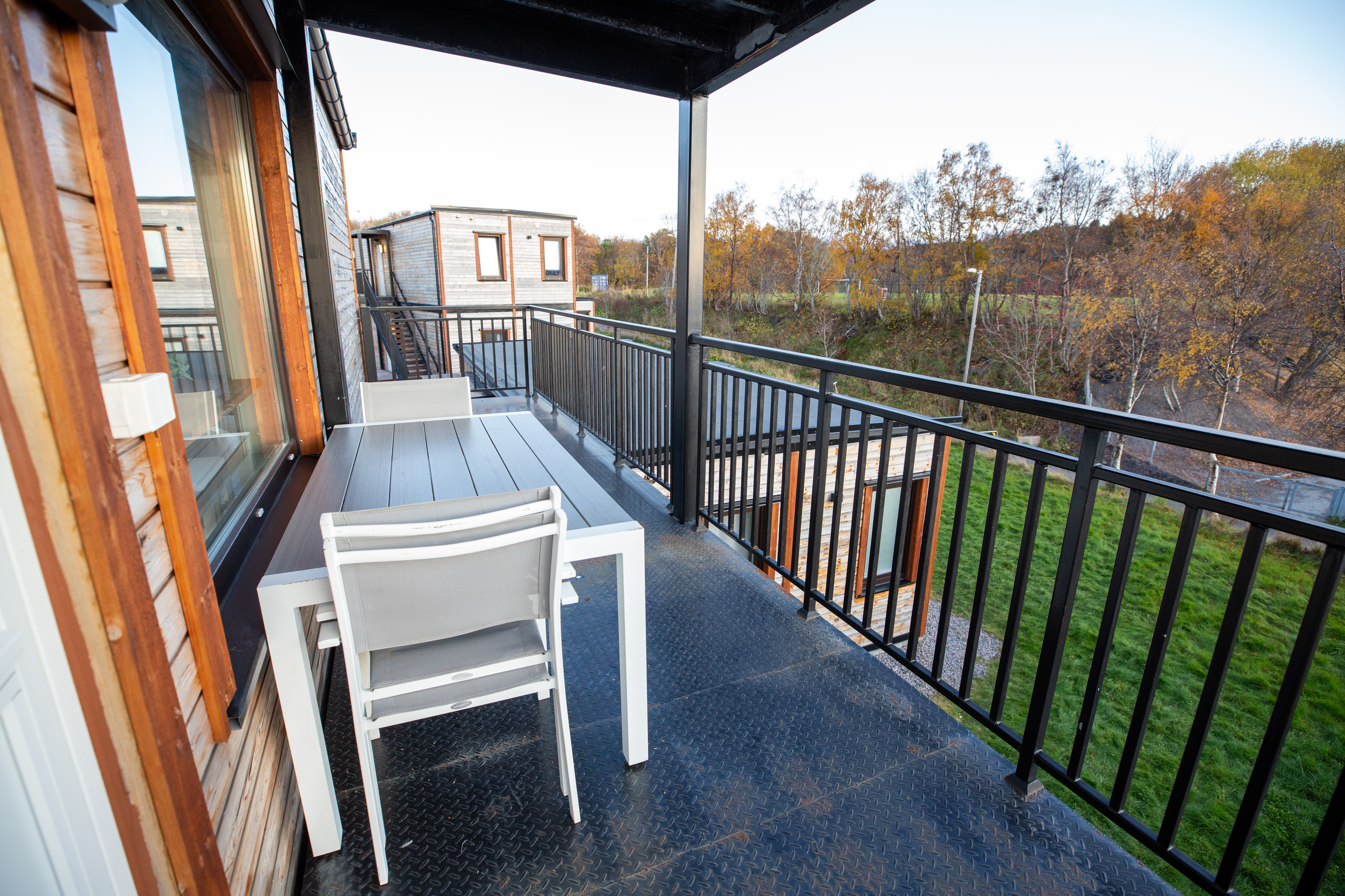 Balkon mit zwei weißen Stühlen und Tisch, Blick auf einen Wohnwagen und Bäume am Campingplatz.