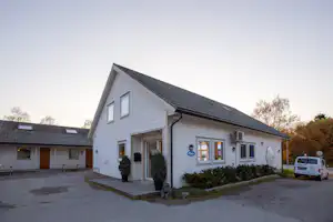 Weißes Camping-Gebäude mit Eingang, Fenstern und Schildern, davor gepflasterter Weg, Hecken und ein geparktes Auto.