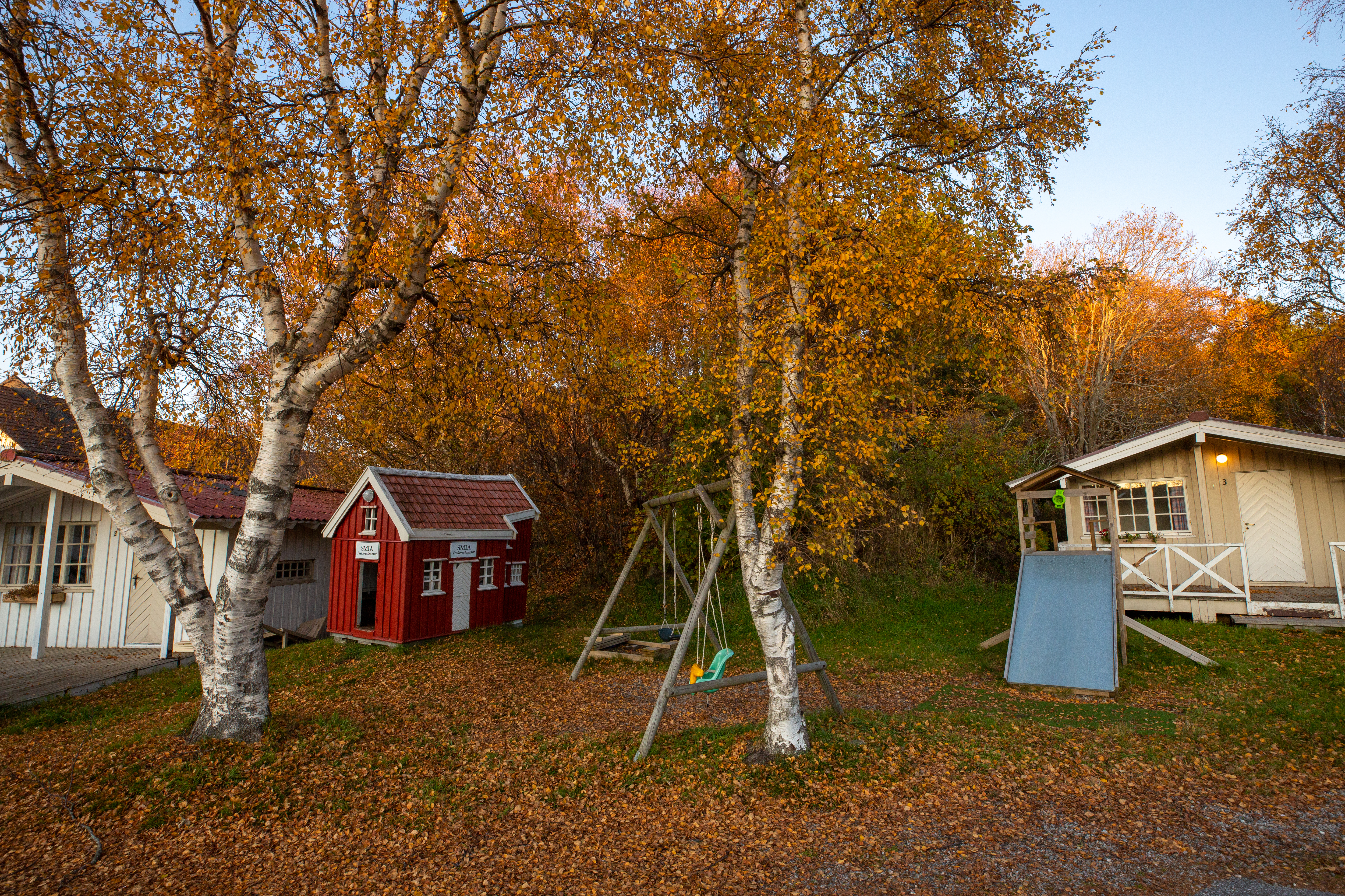 Rote Holzhütte und weißes Gebäude auf einem Campingplatz zwischen Bäumen, mit Schaukeln und Spielgeräten im Vordergrund.