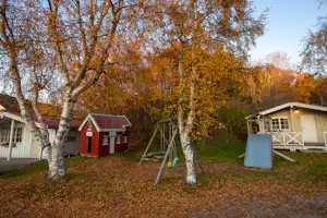 Rote Holzhütte und weißes Gebäude auf einem Campingplatz zwischen Bäumen, mit Schaukeln und Spielgeräten im Vordergrund.