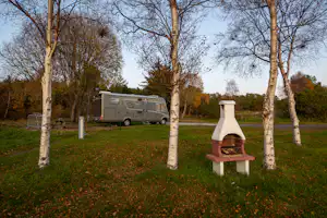 Wohnmobil auf einem Campingplatz zwischen Bäumen, im Vordergrund eine Feuerstelle mit Bank und Grasfläche.