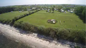 Campingplatz mit grüner Wiese und Wohnwagen am Flussufer, umgeben von Bäumen und kleinen Wegen.