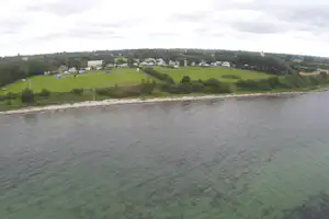 Weite Sicht auf den See mit grünem Ufer und Hügeln, dahinter ein Dorf und Felder unter bewölktem Himmel.