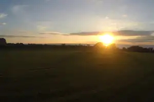 Sonnenuntergang über einer grünen Wiese mit weitem Blick, Feldern und Baumreihen am Horizont.