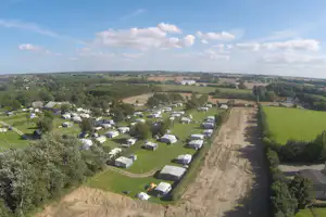 Panoramablick auf einen Campingplatz mit Wohnwagen und Stellplätzen, umgeben von grünen Feldern und einem Weg.