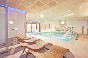 Innenpool mit Wasserbecken, Edelstahl-Liegeflächen und mehreren Liegen im Wellnessbereich, daneben eine Tür zum Ruheraum.