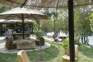 Camping-Pavillon mit Strohdach, Holztischen und Bänken am Flussufer, umgeben von Bäumen und Steinwegen.