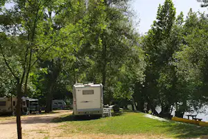Wohnmobil auf einem Stellplatz im Grünen zwischen Bäumen, daneben Campingstühle und ein weiterer Camper.
