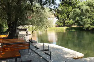 Uferterrasse am Fluss mit Holzbänken, Steintisch und Geländer, umgeben von Bäumen und grünem Wasser.