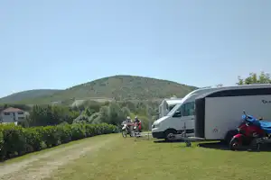 Wohnmobile auf einem Grasstellplatz, Motorräder am Rand, im Hintergrund grüne Hügel und ein Campingplatzweg.