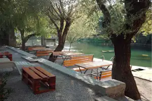 Campingplatz am Fluss mit Sitzbänken und Holztischen am Kiesweg, umgeben von Bäumen und Blick auf das Wasser.