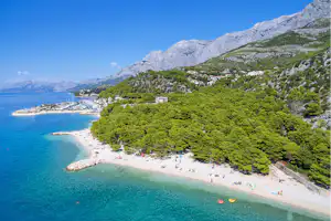 Bucht mit türkisfarbenem Wasser, Sandstrand und vielen Badegästen, umgeben von grünen Hängen und Felsen in den Bergen.