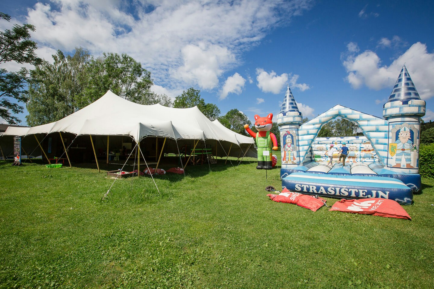 Weiße Zelte auf einer grünen Campingwiese, daneben eine große Hüpfburg mit bunten Motiven.