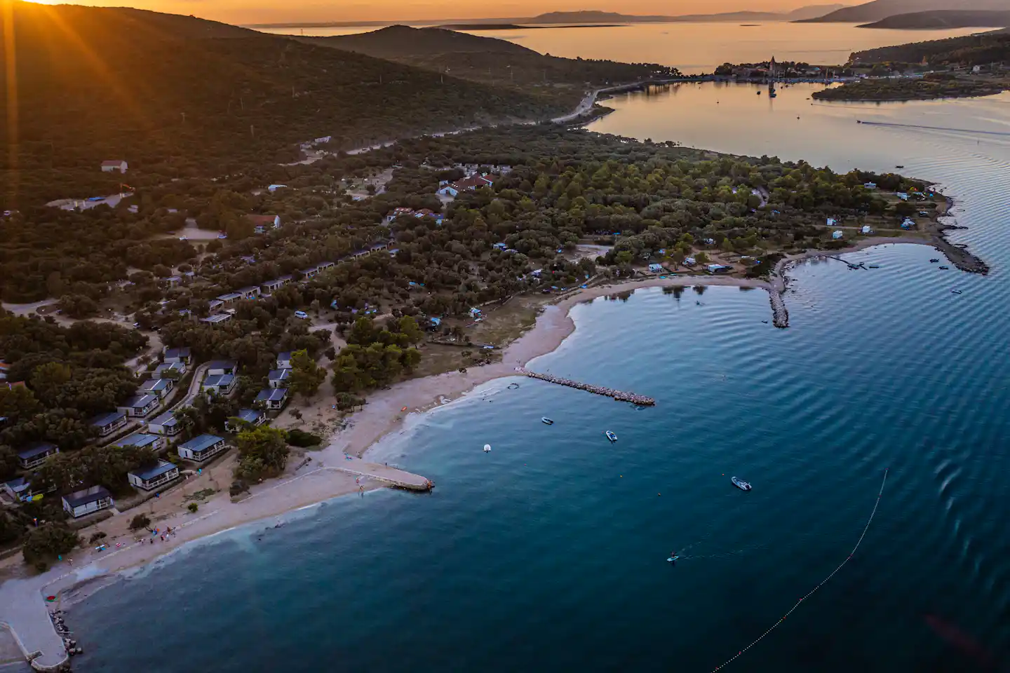 Luftaufnahme eines Campingplatzes am Strand mit Ferienhäusern, Bäumen, Booten und ruhiger Bucht im Abendlicht.