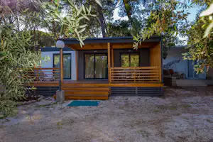 Bungalow mit Holzterrasse und großen Fenstern, umgeben von Bäumen und Sträuchern, mit Sitzbereich am Rand.
