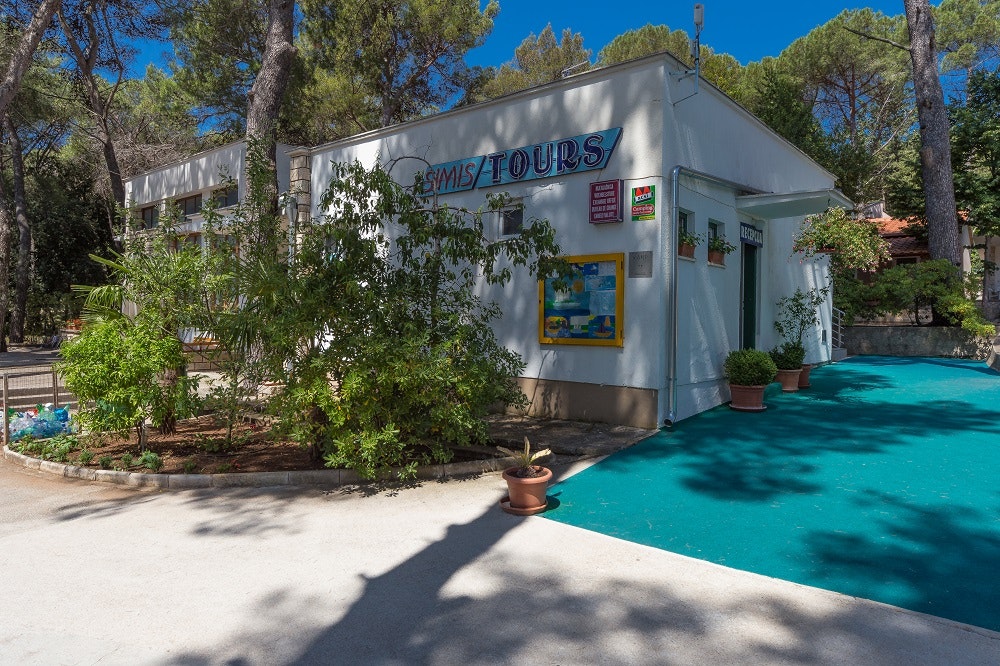 Camping-Unterkunft mit Pool im Vordergrund, weißes Gebäude mit Schildern, umgeben von Bäumen und Sträuchern.