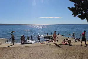 Badeufer am See: Menschen am Strand mit Liegen und Handtüchern, Boote im Wasser, Bäume am Rand.