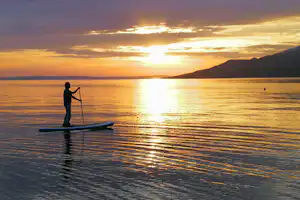 Stand-up-Paddle auf einem See beim Sonnenuntergang, Person auf dem Board mit Paddel, ruhiges Wasser und Berge im Hintergrund.