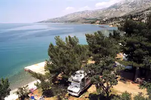 Campingplatz am Meer mit Wohnwagen auf einem Stellplatz, Bäume und Uferpromenade am türkisfarbenen Wasser.