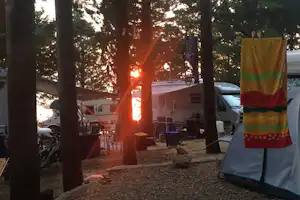 Zelt und Wohnmobil auf dem Campingplatz im Wald, Sonnenuntergang zwischen Bäumen, daneben Campingstühle und Tisch.