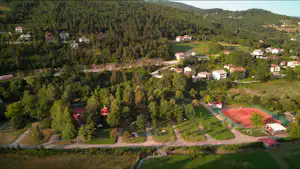 Luftaufnahme eines Campingplatzes mit roten Tennisplätzen, Wegen, Wohnhäusern und viel Wald in hügeliger Landschaft.