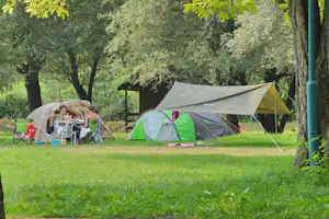 Campingzelt mit grünem Vorzelt und Tisch auf dem Stellplatz im Schatten von Bäumen, daneben weiteres Zelt und Hängematte.