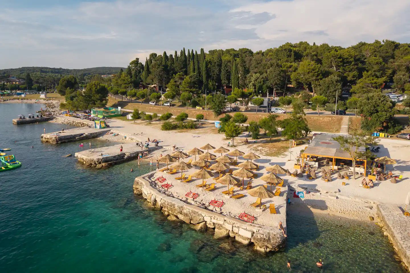 Luftaufnahme eines Campingplatzes am Wasser mit Stegen, Booten und Strandbereich mit Liegen und Sonnenschirmen.