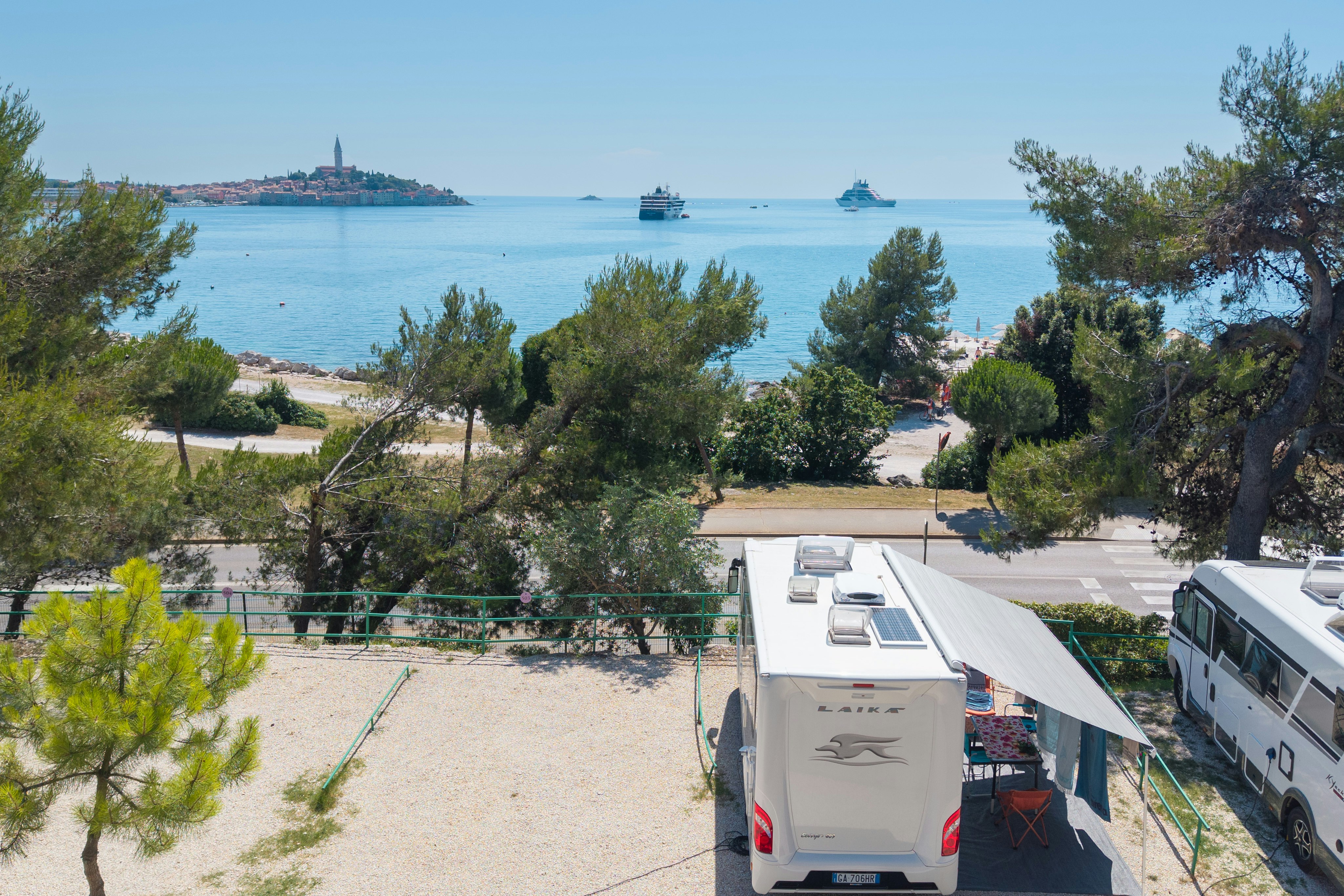 Wohnmobil-Stellplatz am Meer mit Bäumen und Kiesweg, Blick auf Wasser, Boote und eine Insel in der Ferne.