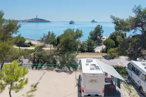 Wohnmobil-Stellplatz am Meer mit Bäumen und Kiesweg, Blick auf Wasser, Boote und eine Insel in der Ferne.