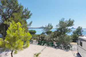 Wohnwagen am Campingplatz mit Kiesfläche, Zaun und Blick auf das Meer zwischen Pinien und Bäumen.