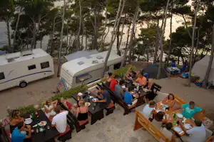 Wohnwagen und Campingtische im Schatten der Bäume, mehrere Personen beim gemeinsamen Essen auf dem Stellplatz.