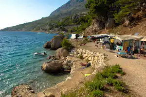 Campingplatz am Meer mit Zelt, Wohnwagen und Campingstühlen am Kiesstrand, Felsen und Berge im Hintergrund.