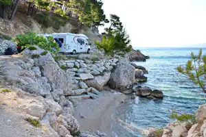 Wohnmobil auf einem Campingplatz am Meer, mit Steinweg, Felsen und Bäumen am Ufer.