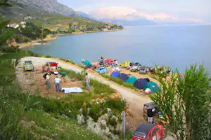 Campingplatz an der Bucht mit Zelten am Ufer, Autos und Wohnwagen entlang des Wegs, im Hintergrund Berge und Meer.