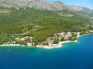 Blick auf einen Campingplatz an der Küste mit Bucht, Strand und Häusern im Grünen unter hohen Bergen.