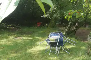 Campingwiese unter Bäumen mit blauem Liegestuhl, rotem Stuhl und grünem Laub im Vordergrund.