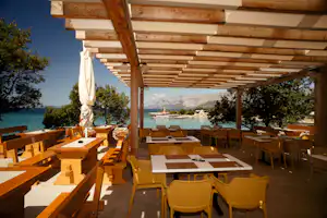 Terrasse mit Holzdach, Tischen und Stühlen am Campingplatz, Blick auf das türkisfarbene Meer und die Berge in der Ferne.