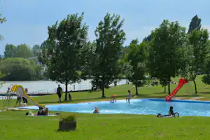 Großer Camping-Pool mit Rutsche und Liegewiese, im Hintergrund Bäume und ein Seeufer mit Geländer.
