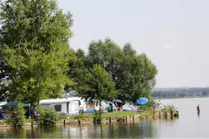 Campingplatz am See: Wohnwagen und Zelte am Ufer hinter einem Holzgeländer, Bäume und ruhiges Wasser im Hintergrund.