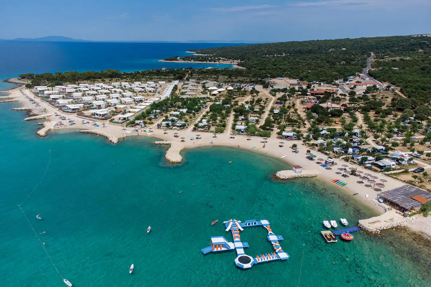 Luftaufnahme eines Campingplatzes am Meer mit Kiesstrand, Booten im Wasser und Reihen von Stellplätzen entlang der Küste.