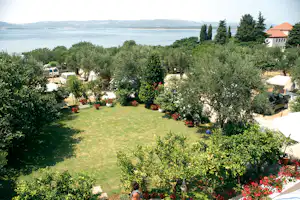 Campinggarten mit Rasenfläche, Olivenbäumen und Blumenbeeten, Wohnwagen und Auto am Rand, Blick auf das Meer.