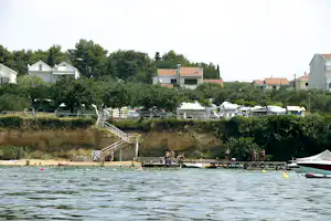 Blick auf den See mit Badebereich, Wohnwagen und Zelten am Ufer, dahinter Häuser und Bäume.